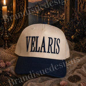 May include: A white and navy blue baseball cap with the word "VELARIS" embroidered in navy blue. The cap has a curved brim and a structured crown. The background includes a candle, a scenic view, and decorative elements.