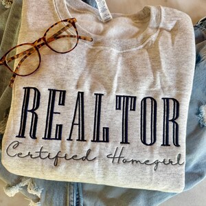 K&ouml;nnte beinhalten: Ein graues Sweatshirt mit dem gestickten Text "REALTOR Certified Homegirl" auf der Vorderseite. Das Sweatshirt ist gefaltet und liegt auf einer blauen Jeans.