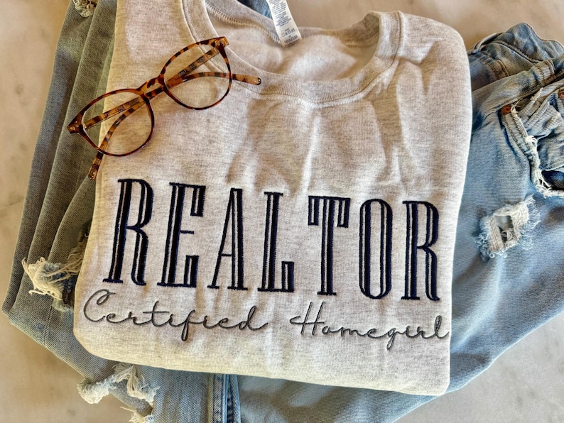 K&ouml;nnte beinhalten: Ein graues Sweatshirt mit dem gestickten Text "REALTOR Certified Homegirl" auf der Vorderseite. Das Sweatshirt ist gefaltet und liegt auf einer blauen Jeans.