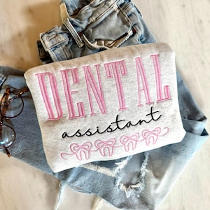 May include: A light grey sweatshirt embroidered with the words "DENTAL assistant" in pink and black, with pink tooth designs below. It rests on light blue jeans and a pair of spectacles.