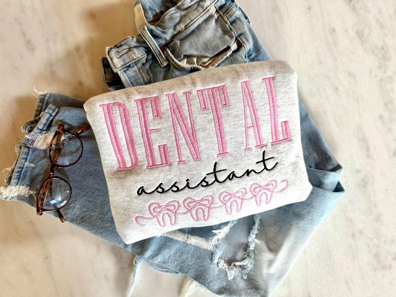 May include: A light grey sweatshirt embroidered with the words "DENTAL assistant" in pink and black, with pink tooth designs below. It rests on light blue jeans and a pair of spectacles.