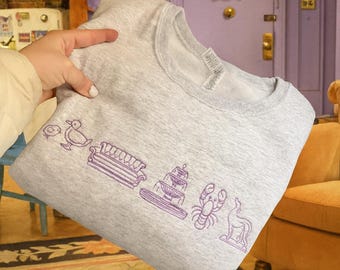 May include: A grey sweatshirt featuring purple embroidered icons of a duck, sofa, cake, lobster, and dog. The sweatshirt is held up in front of a purple door and yellow sofa.
