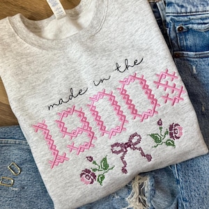 May include: A heather gray sweatshirt with the phrase "made in the 1900s" embroidered in pink. The design includes a bow and floral accents. The sweatshirt is displayed on a pair of blue jeans.