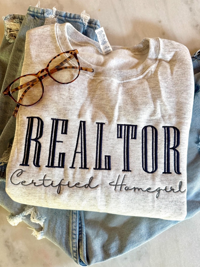 K&ouml;nnte beinhalten: Ein graues Sweatshirt mit dem Text "REALTOR Certified Homegirl" in schwarzer Stickerei.