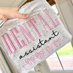 May include: A pale grey sweatshirt featuring the embroidered words "DENTAL assistant" in pink and black. Below the text are pink bow-tied teeth. The sweatshirt is held up by a hand.