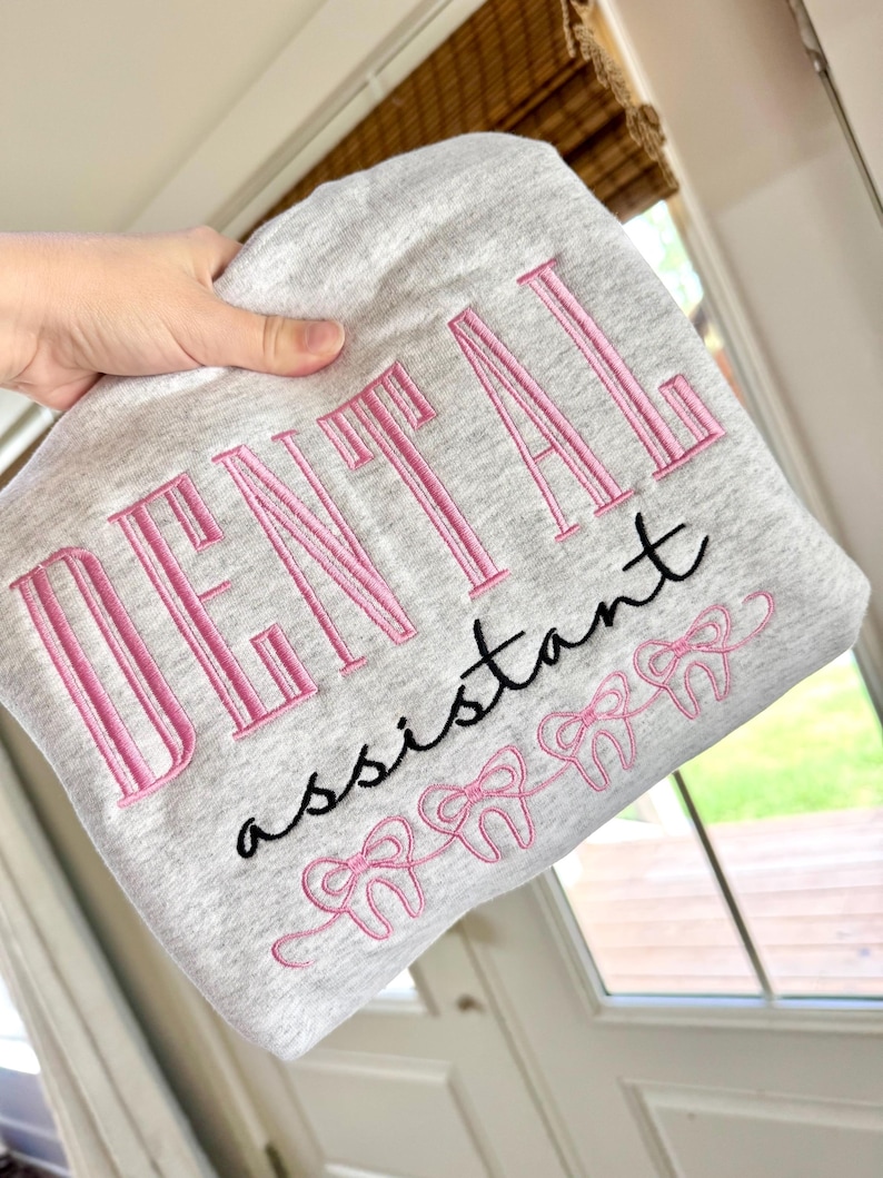 May include: A pale grey sweatshirt featuring the embroidered words "DENTAL assistant" in pink and black. Below the text are pink bow-tied teeth. The sweatshirt is held up by a hand.