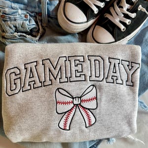 May include: A gray sweatshirt with the text "GAMEDAY" in black outlined letters and a white baseball-themed bow with red stitching.