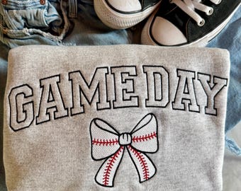 May include: A gray sweatshirt with the text "GAMEDAY" in black outlined letters and a white baseball-themed bow with red stitching.