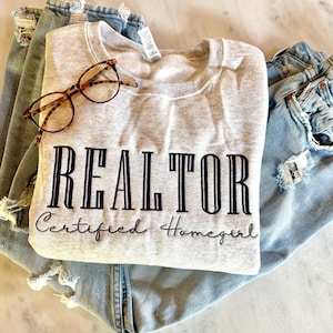 May include: A gray sweatshirt with the text "REALTOR Certified Homegirl" in black lettering. The sweatshirt is folded and lying on top of a pair of blue jeans.