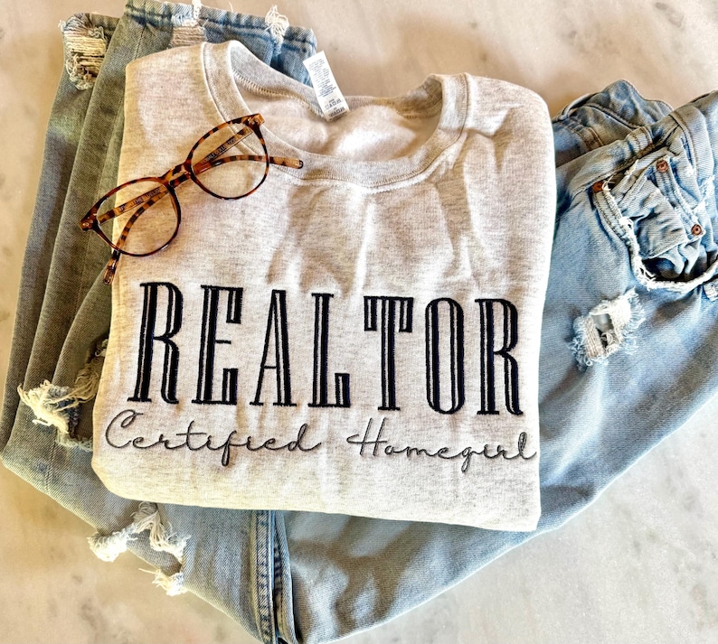 K&ouml;nnte beinhalten: Ein graues Sweatshirt mit dem Text "REALTOR Certified Homegirl" in schwarzen Buchstaben. Das Sweatshirt ist gefaltet und liegt auf einer blauen Jeans.