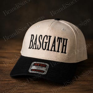 以下が含まれることがあります： ベージュと黒の野球帽。黒で刺繍された「BASGIATH」の文字が特徴です。つばは黒で、上部に黒いボタンが付いています。「OTTO」のタグが付いています。