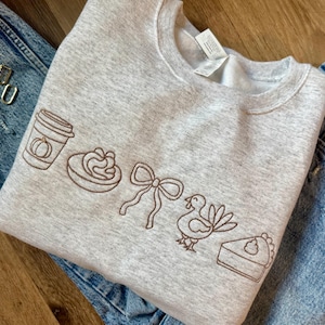 May include: A light gray sweatshirt with embroidered brown line art of a pumpkin spice latte, a swirl of whipped cream, a bow, a turkey, and a slice of pie. The sweatshirt is folded on top of a pair of blue jeans.