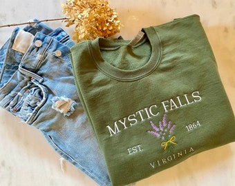 May include: A green sweatshirt with the text "MYSTIC FALLS EST. 1864 VIRGINIA" embroidered on the front. The text is in white with a purple and yellow floral design.