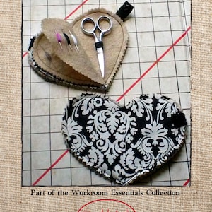 May include: A heart-shaped needle book with a black and white damask pattern. The book is open to reveal a smaller heart-shaped compartment with a pair of scissors and needles. The text "The Nifty Needle Book" is at the top of the image. The text "Part of the Workroom Essentials Collection" is below the image. The text "red twig DIY" is at the bottom of the image.
