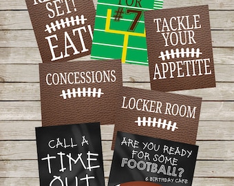 May include: Eight printable signs with a football theme. The signs have white text on a variety of backgrounds, including brown leather, green, black, and chalkboard. The signs include phrases like "Ready! Set! Eat!" and "Tackle Your Appetite".