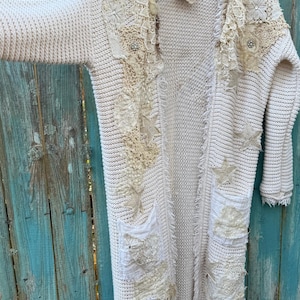 May include: Cream-colored, long open cardigan sweater with decorative patches of lace, stars, and embellishments. The sweater has two front pockets and a frayed hem. The background is a weathered, turquoise wooden fence.