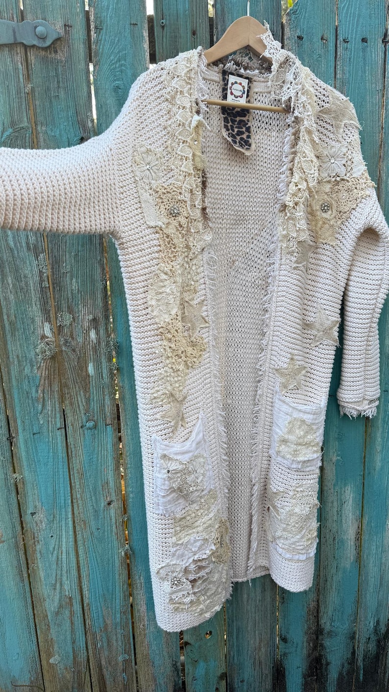 May include: Cream-colored, long open cardigan sweater with decorative patches of lace, stars, and embellishments. The sweater has two front pockets and a frayed hem. The background is a weathered, turquoise wooden fence.
