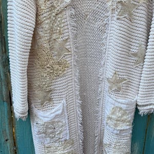 May include: Cream-colored, open-front cardigan sweater with intricate embellishments. Features star-shaped appliqu&eacute;s, lace detailing, and pockets. The sweater has a textured knit design and a relaxed fit, perfect for layering.