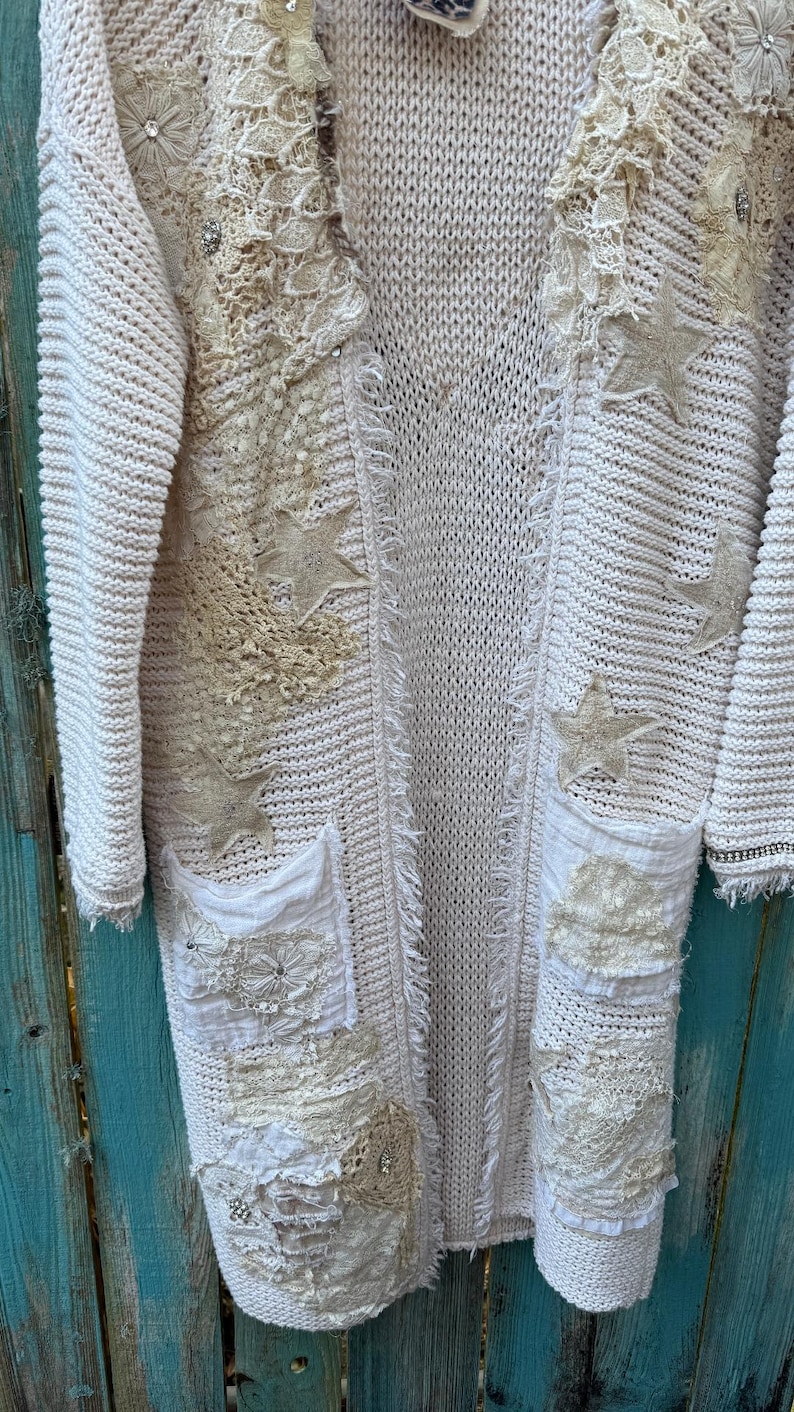 May include: Cream-colored, open-front cardigan sweater with intricate embellishments. Features star-shaped appliqu&eacute;s, lace detailing, and pockets. The sweater has a textured knit design and a relaxed fit, perfect for layering.