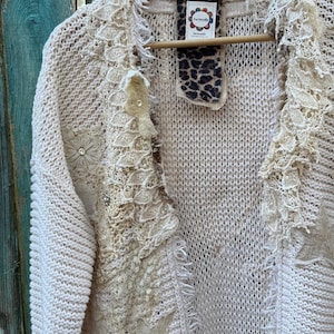 May include: Cream-colored, open-front cardigan sweater with intricate lace and star appliqu&eacute;s. The sweater features a V-neck, fringe detailing, and is embellished with floral and rhinestone accents. It hangs on a wooden hanger against a weathered blue background.