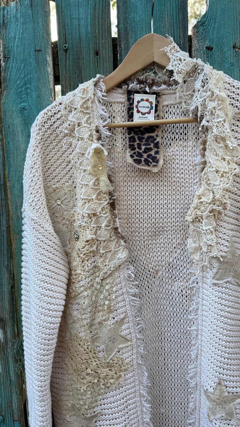May include: Cream-colored, open-front cardigan sweater with intricate lace and star appliqu&eacute;s. The sweater features a V-neck, fringe detailing, and is embellished with floral and rhinestone accents. It hangs on a wooden hanger against a weathered blue background.