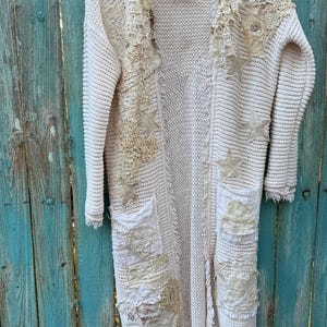 May include: Cream-colored, long cardigan sweater with decorative lace and star appliqu&eacute;s. The open-front sweater features two front pockets and is hanging on a wooden hanger. The sweater is made of a chunky knit material and has a distressed, bohemian style.