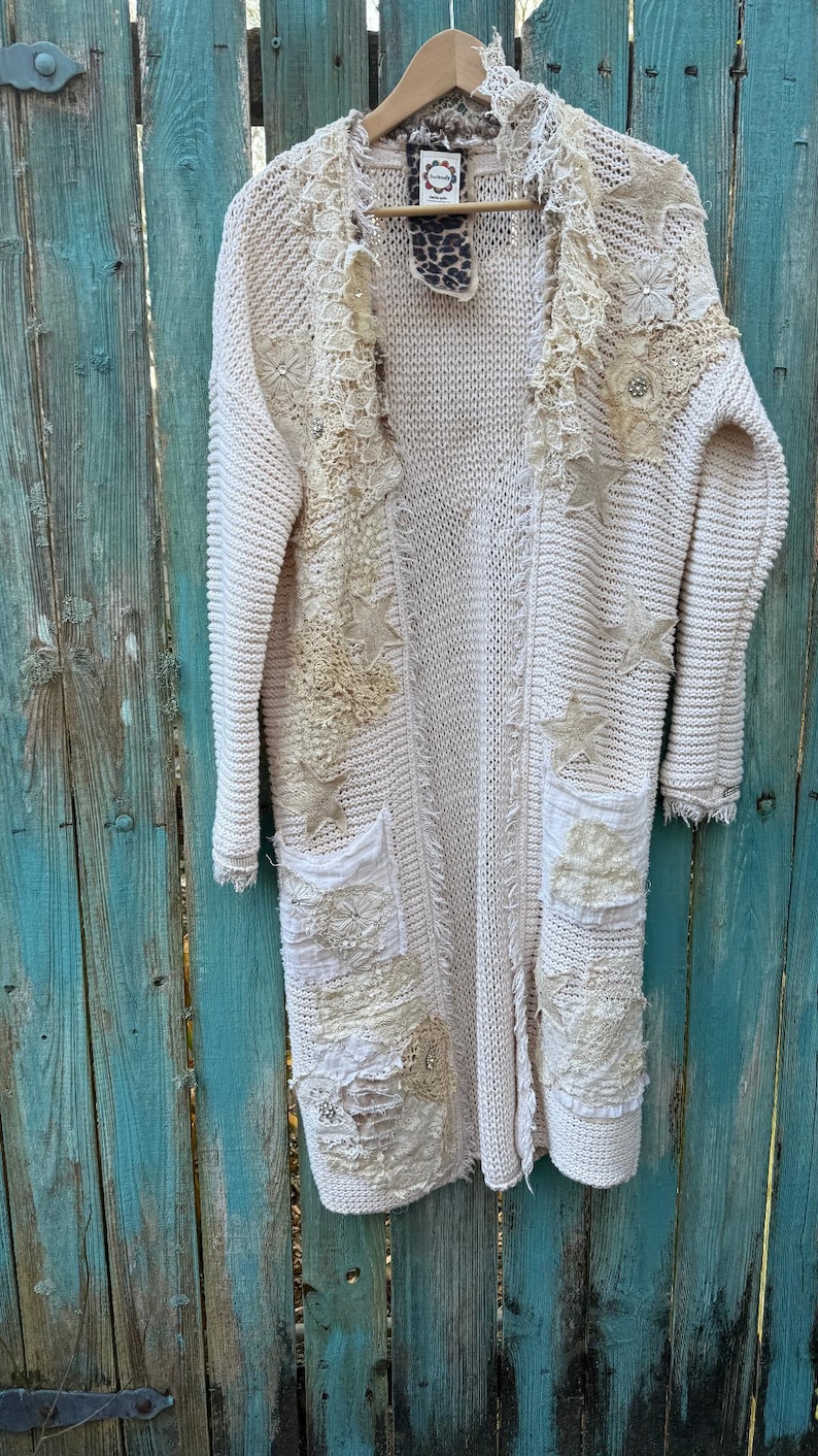 May include: Cream-colored, long cardigan sweater with decorative lace and star appliqu&eacute;s. The open-front sweater features two front pockets and is hanging on a wooden hanger. The sweater is made of a chunky knit material and has a distressed, bohemian style.