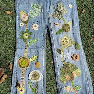 May include: Light blue denim jeans with colorful floral and leaf appliqués. The jeans feature a flared leg and are decorated with various fabric patches in shades of green, yellow, and brown. The jeans are lying on a bed of green grass.