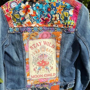 May include: A blue denim jacket with colorful floral patchwork on the upper back and shoulders. The back features a rectangular patch with the words "STAY WILD MOON CHILD" and a mushroom graphic. The jacket has a vintage, bohemian aesthetic.