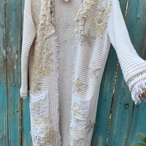 May include: Cream-colored, open-front cardigan with intricate lace and star appliqu&eacute;s. The long sweater features pockets and fringed edges, creating a bohemian aesthetic. The cardigan is hanging on a wooden hanger.