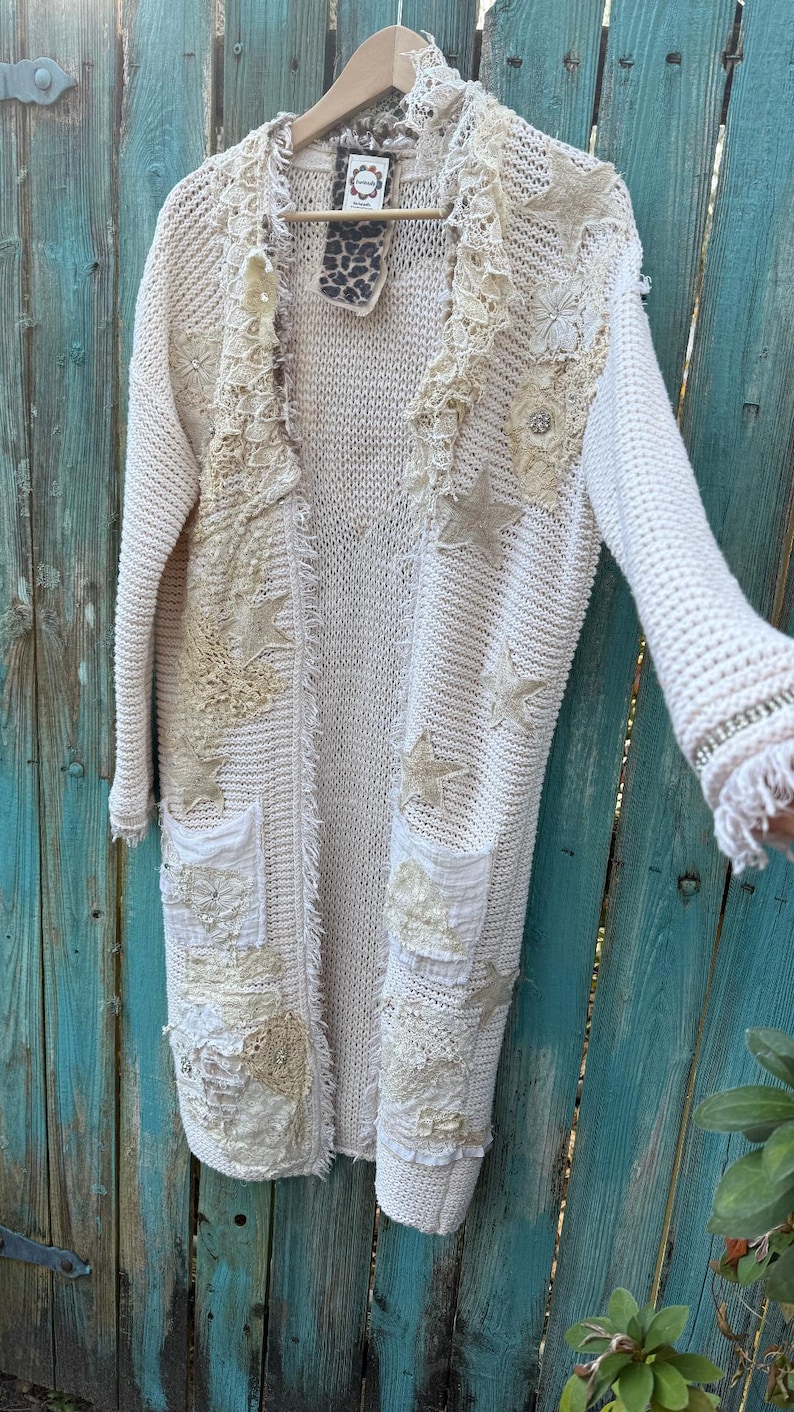 May include: Cream-colored, open-front cardigan with intricate lace and star appliqu&eacute;s. The long sweater features pockets and fringed edges, creating a bohemian aesthetic. The cardigan is hanging on a wooden hanger.