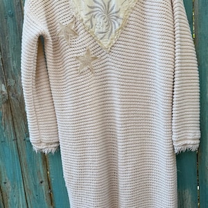May include: Cream-colored, long-sleeved cardigan with a textured knit design. The cardigan features a decorative heart-shaped patch on the back, embellished with floral embroidery and two star-shaped appliqu&eacute;s. The sleeves have a fringed hem.