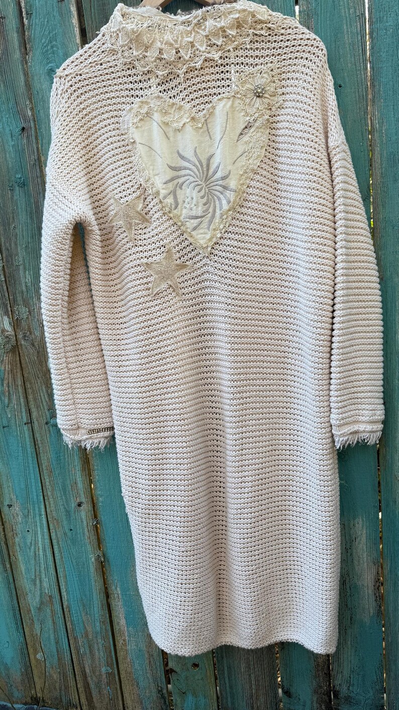May include: Cream-colored, long-sleeved cardigan with a textured knit design. The cardigan features a decorative heart-shaped patch on the back, embellished with floral embroidery and two star-shaped appliqu&eacute;s. The sleeves have a fringed hem.