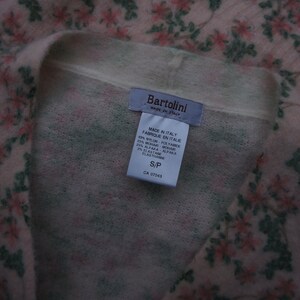 Może przedstawiać: R&oacute;żowo-zielony kardigan w kwiaty z białą metką, na kt&oacute;rej widnieje napis "Bartolini Made in Italy" oraz skład tkaniny: 49% nylon, 25% moher, 25% alpaka, 1% elastan. Metka zawiera r&oacute;wnież informację o rozmiarze S/P i numerze CA 57043.