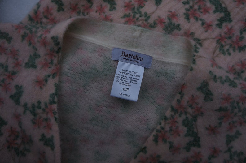 Może przedstawiać: R&oacute;żowo-zielony kardigan w kwiaty z białą metką, na kt&oacute;rej widnieje napis "Bartolini Made in Italy" oraz skład tkaniny: 49% nylon, 25% moher, 25% alpaka, 1% elastan. Metka zawiera r&oacute;wnież informację o rozmiarze S/P i numerze CA 57043.