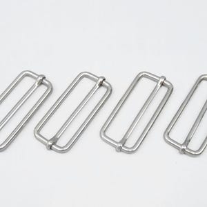 May include: Four silver-colored rectangular metal slides, each with a central bar. The slides are arranged diagonally against a white background. These hardware components are likely used in various crafting and sewing projects.