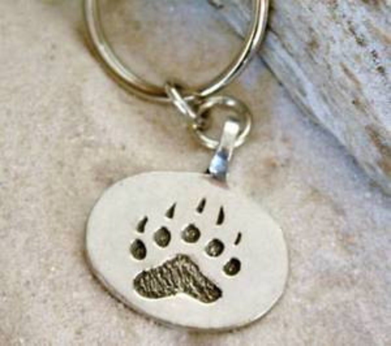 K&ouml;nnte beinhalten: Silberner ovaler Schl&uuml;sselanh&auml;nger mit einem B&auml;renpfotenabdruck.