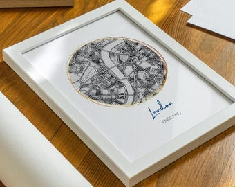 Può includere: Una stampa incorniciata bianca con una mappa in bianco e nero di Londra, Inghilterra. La mappa è circondata da un cerchio dorato e il testo "Londra, Inghilterra" è scritto in corsivo blu.