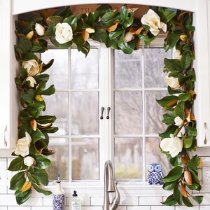 May include: A magnolia garland with large green leaves, brown accents, and creamy white flowers frames a white window. The garland is draped around the window frame, creating a natural and elegant decorative element. The kitchen window also features a small blue and white owl figurine.