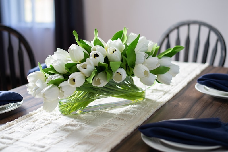 May include: A centerpiece of white tulips in a clear glass vase sits on a cream-colored textured table runner. The arrangement is on a dark wooden table, with navy blue napkins and plates visible.