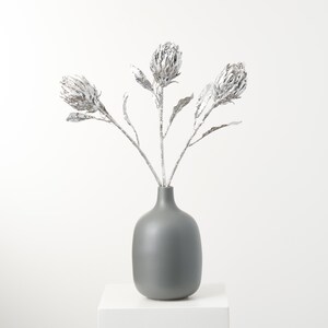 May include: A bouquet of three silver protea flowers in a gray ceramic vase. The vase is on a white pedestal.