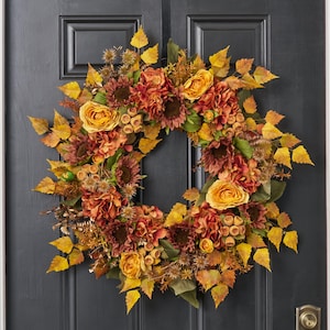 May include: A fall-themed wreath with a variety of flowers and foliage. The wreath features sunflowers, roses, and hydrangeas in shades of orange, yellow, and brown, with accents of green leaves and berries. The wreath is displayed on a dark gray door.