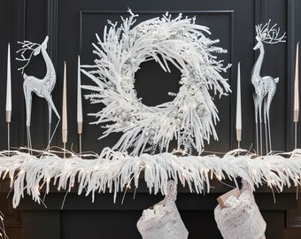 Op de afbeelding: Een witte, met sneeuw bedekte kerstkrans met zilveren accenten, twee zilveren rendierfiguren en een witte, met sneeuw bedekte guirlande met lampjes op een zwarte schouw. Twee witte nep bontkousen hangen onder de schouw.