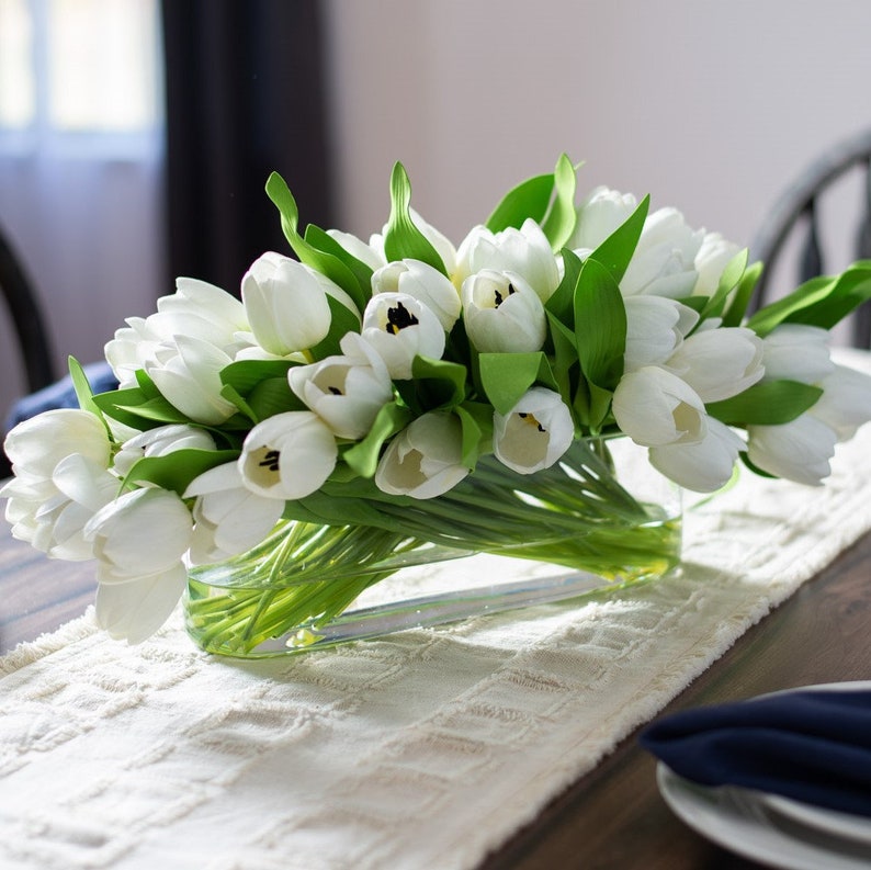 May include: A rectangular glass vase filled with white tulips and green leaves. The arrangement sits on a cream-colored table runner. The table is set with a dark blue napkin and white plates.