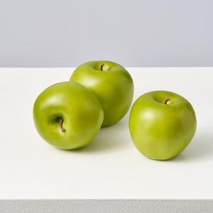 May include: Three green apples sit on a white surface. Each apple has a smooth, shiny skin and a small brown stem. The apples are a vibrant green colour, and the lighting creates highlights and shadows.