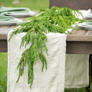 May include: A white table runner with frayed edges is decorated with a green garland of pine sprigs. The runner is laid on a wooden table with two place settings on each side.