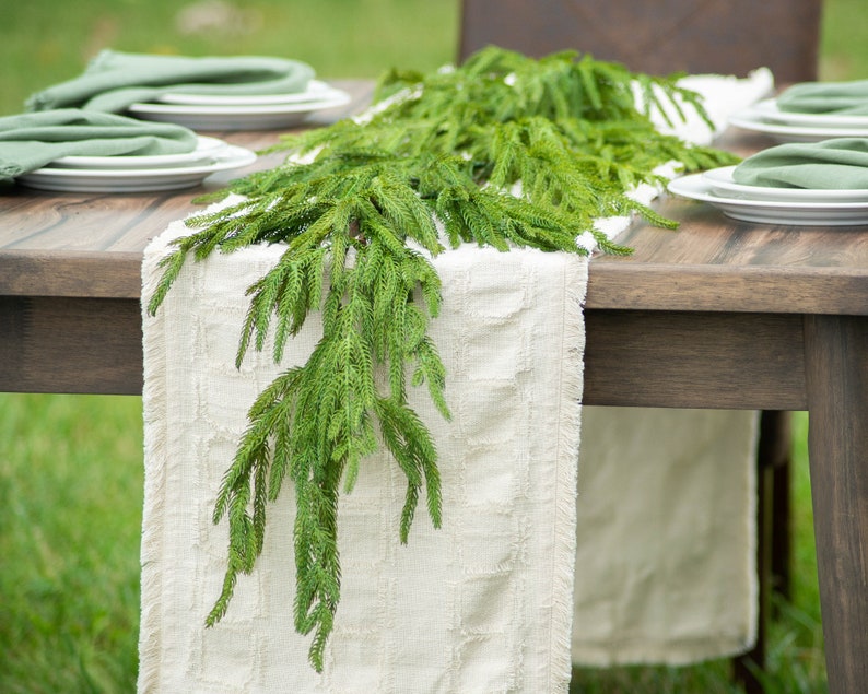 May include: A white table runner with frayed edges is decorated with a green garland of pine sprigs. The runner is laid on a wooden table with two place settings on each side.