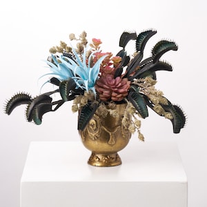 May include: A gold-colored urn with embossed skull and drape details holds an arrangement of artificial plants, including dark green Venus flytraps, light blue spiky succulents, pink rosette succulents, and small beige dried floral sprigs.