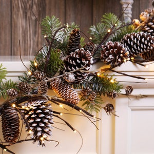 Puede incluir: Una guirnalda de ramas marrones con ramas de pino y piñas artificiales decoradas con nieve artificial blanca. La guirnalda está adornada con luces de hadas blancas cálidas.