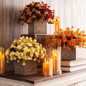 May include: Three wooden boxes with different colored artificial flower arrangements and lit candles. The boxes are stacked on top of each other on a white wooden surface.